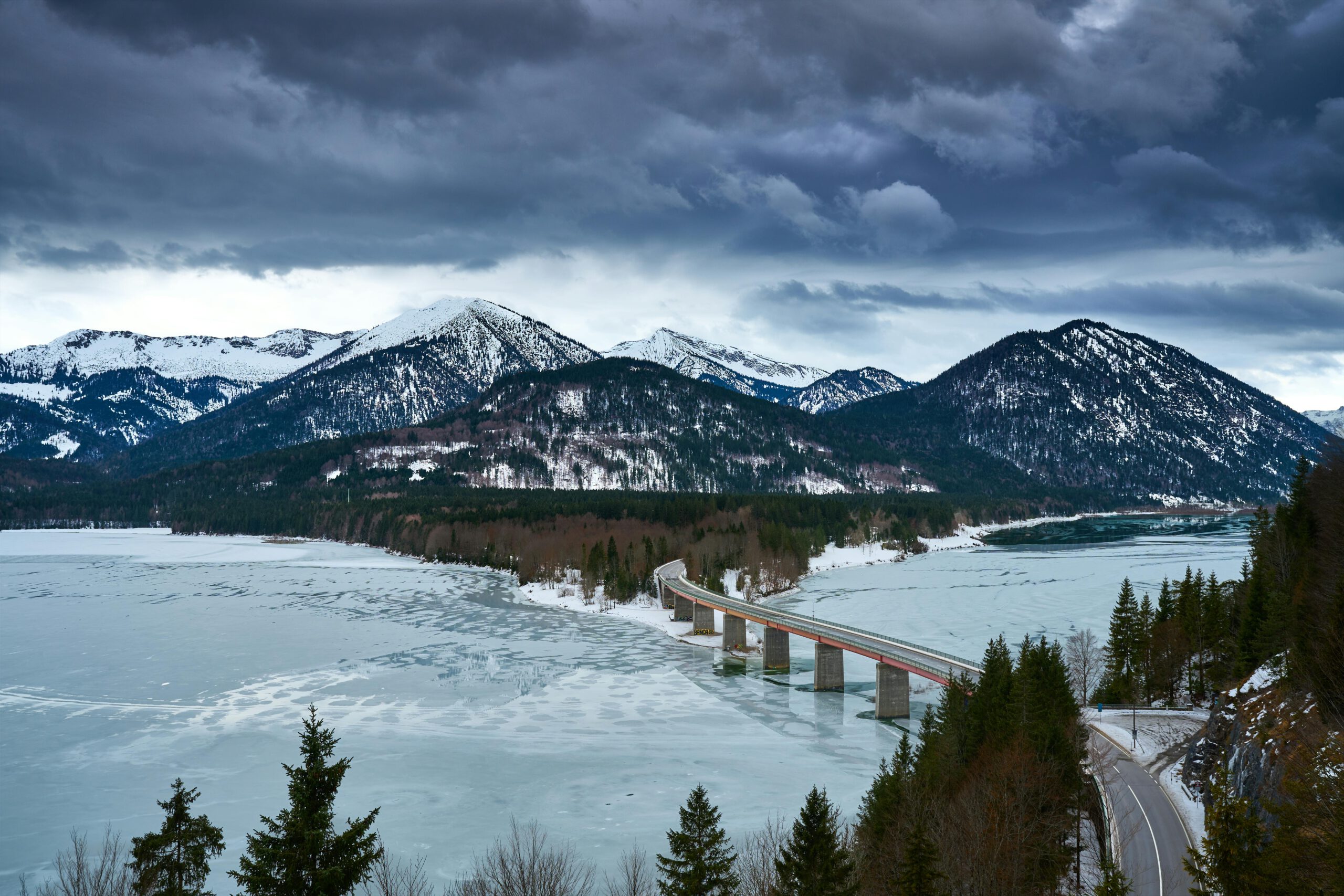 Atemberaubender Winterblick auf die Brücke über den zugefrorenen See und die schneebedeckten bayerischen Alpen in Lenggries, Deutschland.