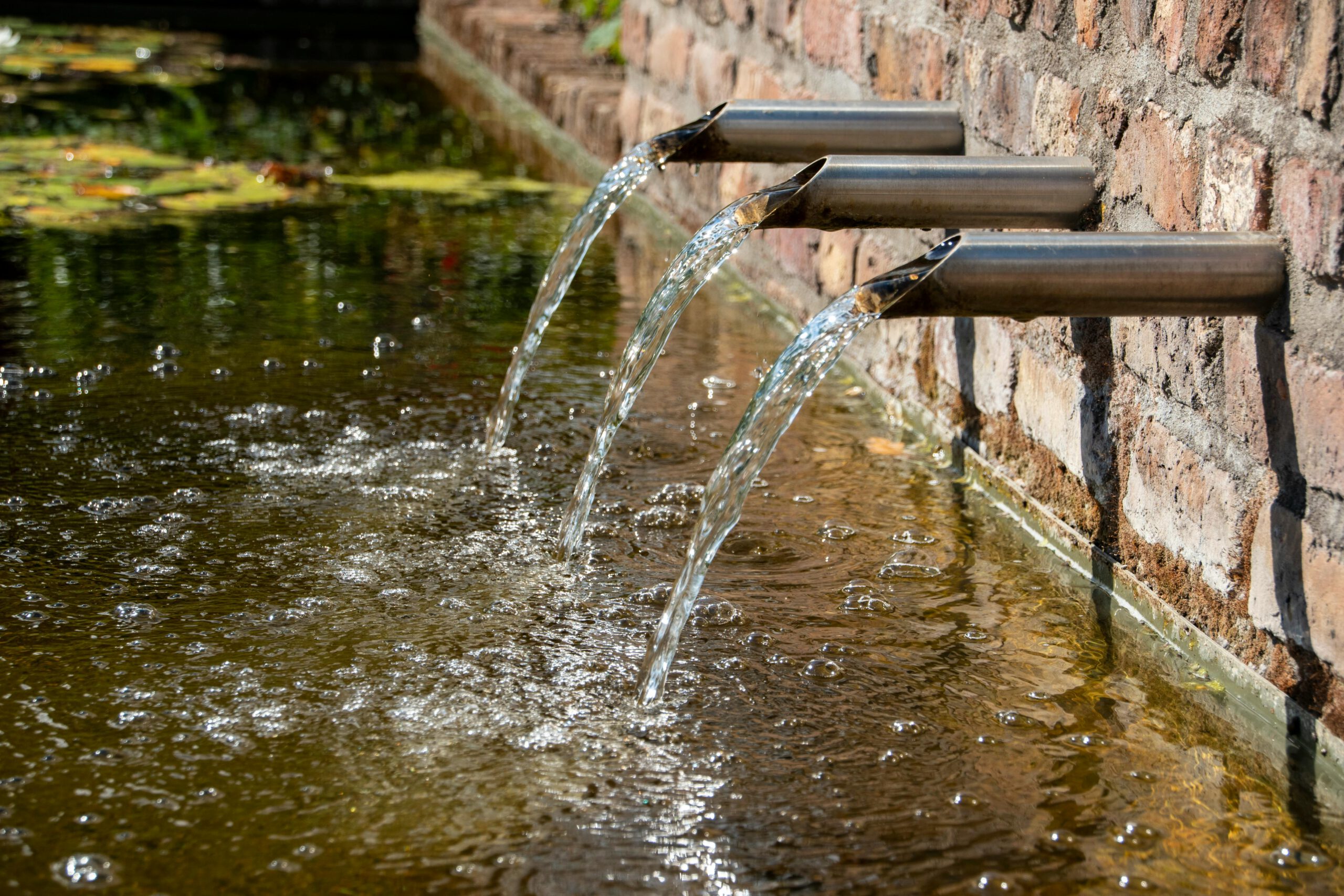 Kristallklares Wasser ergießt sich aus einem modernen Edelstahlbrunnen in einen idyllischen Gartenteich.