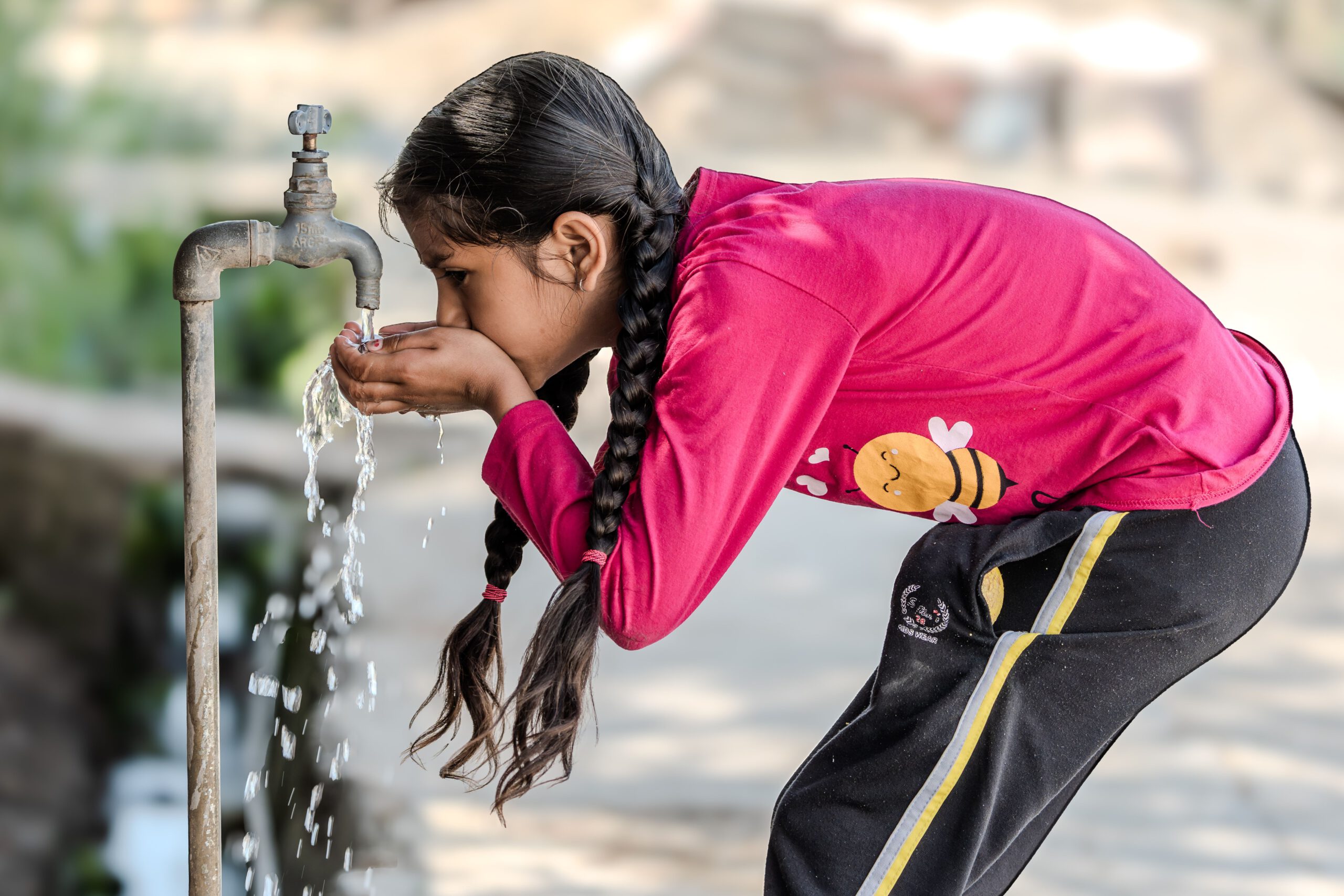 Ein junges Mädchen trinkt an einem sonnigen Tag Wasser aus einem Wasserhahn im Freien und unterstreicht damit die Einfachheit des ländlichen Lebens.
